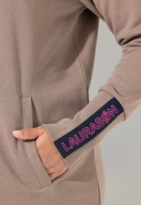 Vaaleanruskea huppari, jossa on pitkät hihat. Hupparissa on tummansininen käsivarsi, jossa on vaaleanpunaista tekstiä "LAURASON". Mukana on sivutasku.