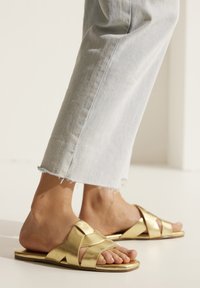 Gold slide sandals feature a crisscross strap design. The sole is flat and smooth, complemented by tapered toes. Worn with light blue jeans.