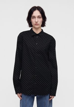 Personne aux cheveux courts et foncés portant une chemise noire ample à motifs et un jean bleu, debout devant un fond clair uni.