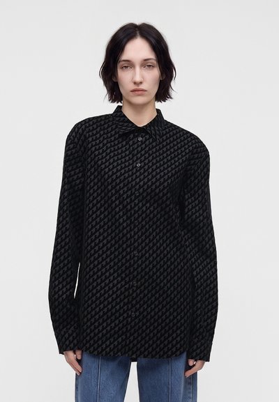 Personne aux cheveux courts et foncés portant une chemise noire ample à motifs et un jean bleu, debout devant un fond clair uni.