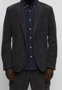 Blazer in velluto a coste grigio con colletto a scollatura, tasche frontali e una tasca sul petto, indossato sopra una camicia blu navy con chiusura a bottoni.