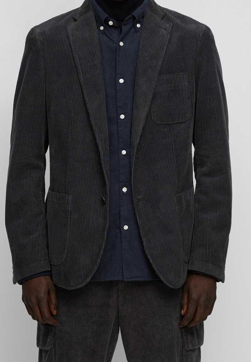 Blazer in velluto a coste grigio con colletto a scollatura, tasche frontali e una tasca sul petto, indossato sopra una camicia blu navy con chiusura a bottoni.