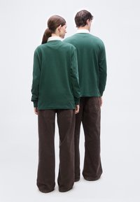 Δύο άτομα στέκονται πλάτη με πλάτη, φορώντας πράσινα μακρυμάνικα μπλουζάκια πάνω από λευκά γιακάδες και φαρδιά καφέ παντελόνια σε ανοιχτό φόντο.