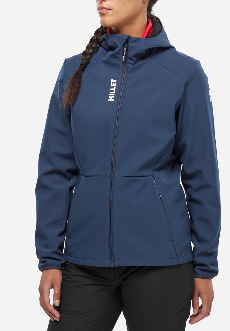 Veste softshell navy avec fermeture éclair sur le devant, capuche et poignets élastiques. Présente un intérieur rouge contrastant et un logo de marque sur la poitrine.