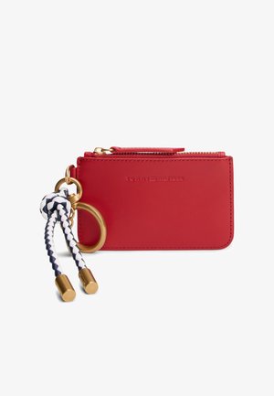 Cartera de piel roja con herrajes dorados, cierre de cremallera y llavero trenzado en azul marino y blanco. Presenta un logotipo de la marca en relieve.