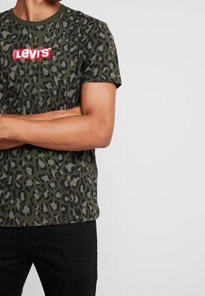 Homme portant un t-shirt Levi's imprimé léopard vert et noir, les bras croisés, associé à un pantalon noir, sur un fond uni.