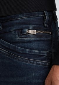 Jeans de mezclilla oscura que presentan un primer plano de un bolsillo lateral con cremallera metálica y tejido texturizado, mostrando detalles de costura.