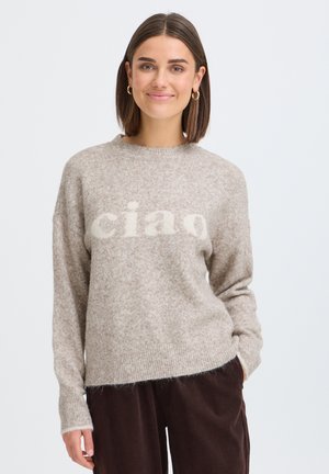 Femme aux cheveux courts bruns portant des boucles d'oreilles créoles dorées, un pull beige avec le texte "ciao" et un pantalon marron foncé, souriante, la main dans la poche.