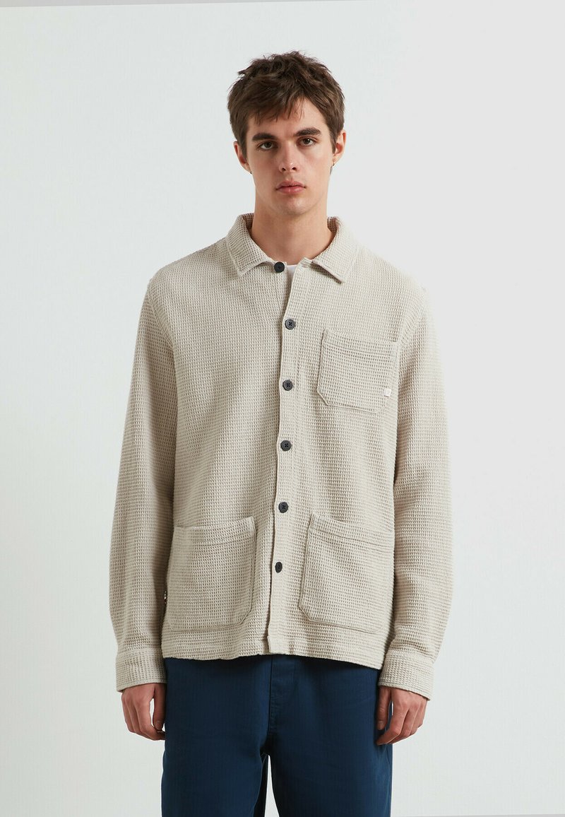 Giovane uomo che indossa un cardigan beige a trama con bottoni e tre tasche frontali, abbinato a pantaloni blu scuro, in piedi di fronte a uno sfondo neutro.