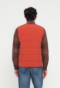 Κόκκινο καπιτονέ γιλέκο με ψηλό γιακά, που αποκαλύπτει μια ομαλή υφή. Φοριέται πάνω από ένα καρό πουκάμισο με μακριά μανίκια και μπλε τζιν.