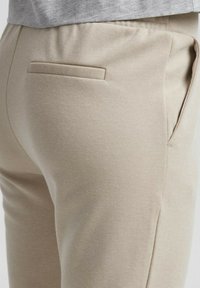 Beige broek gemaakt van getextureerde stof, met een enkele achterzak en zijzakken. De tailleband is elastisch voor comfort.