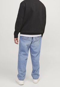 Uomo che indossa jeans azzurri chiari, sneaker bianche, felpa nera e maglietta bianca, in piedi di spalle contro uno sfondo neutro.