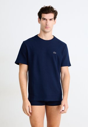 Navy blue short-sleeve T-shirt with a round neck, textured fabric, and a small embroidered logo on the chest, paired with dark shorts.