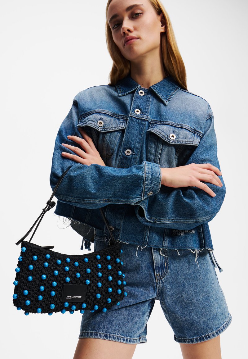 Svart virkat handväsk med blå pärlor, med ett kort handtag, hålls mot en bakgrund av denimjacka och shorts. Rent, modernt design.