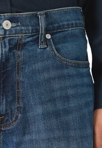 Jeans din denim de un albastru închis, cu un design clasic cu cinci buzunare, cusături de contrast, fermoar metalic și o textură subtilă.