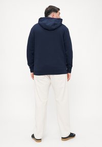 Felpa blu navy con cappuccio regolabile, vestibilità comoda, polsini e orlo a coste, abbinata a pantaloni beige larghi e sandali neri.