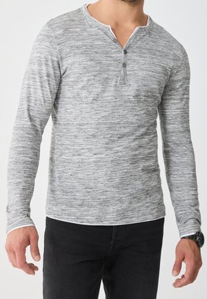 Homme portant un t-shirt henley gris chiné à manches longues avec trois boutons et un pantalon noir, debout les bras détendus.