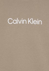 Beżowy t-shirt o gładkiej fakturze z napisem "Calvin Klein" wydrukowanym na piersiach dużymi białymi literami. Prosty i minimalistyczny design.