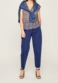 Blouse à motifs floraux avec des manches volantes, associée à un pantalon bleu marine ajusté. Une veste en denim est drapée sur les épaules. Talons en imprimé serpent.