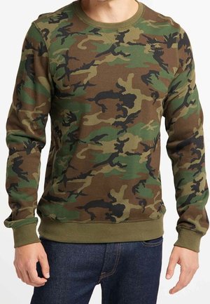 Homme portant un sweat-shirt camouflage vert et marron à manches longues et un jean bleu, debout les bras relâchés le long du corps.