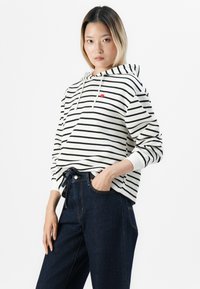 Jeune femme portant un sweat à capuche blanc et noir rayé avec un petit cœur rouge et un jean taille haute bleu foncé, se tenant avec une main dans la poche.