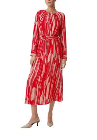 Femme portant une robe midi à manches longues avec un motif abstrait rouge et beige, assortie d'une ceinture et de talons slingback beiges à bout pointu.