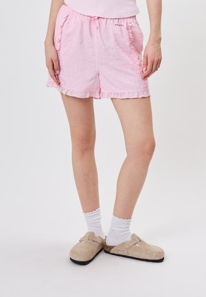 Person iført lyserøde flæseshorts, hvide ankelsokker og beige slip-on træsko, stående på hvid baggrund.