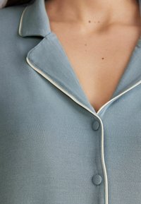 Lichtblauw pyjamaje met een zachte textuur, voorzien van een knoopsluiting aan de voorkant, een opstaande kraag en witte piping.
