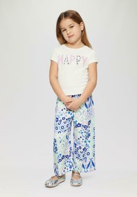Witte t-shirt met "HAPPY" in roze en paarse pailletten, gecombineerd met losse broek in bloemenprint in blauw en groen, en zilveren schoenen.