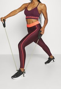 Kobieta w maroonowym odzież sportowej i czarnych sneakersach ćwiczy z taśmami oporowymi w pozycji wykroku, na jednolitym tle.