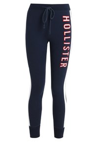 Joggers de color navy con paneles laterales blancos, que cuentan con una cintura ajustable con cordón y la palabra "HOLLISTER" en letras rojas en negrita a lo largo de la pierna izquierda.