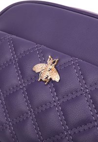 Borsa in pelle trapuntata viola con un'applicazione dorata a forma di ape, con dettagli di cucitura visibili e una texture liscia.