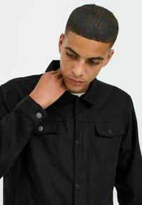 Veste en daim noir avec un col, deux poches poitrine et des fermetures à boutons-pression. La texture semble douce et mate.