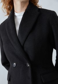 Zwarte blazer van een wolmix met een dubbelbreasted ontwerp, knopenrevers en twee voorzakken met knoopaccenten. Zachte textuur.