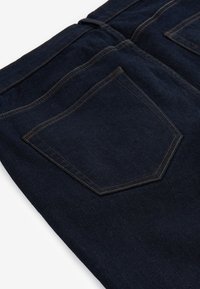 Donkerblauwe denim jeans die de achterzak en stiksel details laten zien op een witte achtergrond.