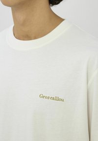 T-shirt en coton blanc à coupe décontractée, avec un texte brodé vert « Gros caillou » sur le côté gauche de la poitrine.