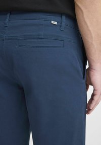 Pantaloni bleu marin dintr-un material ușor, având o buzunar la spate cu o cusătură ascunsă și un mic eticheta a brandului pe talie.