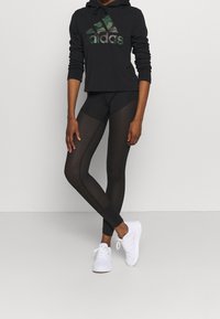 Sudadera corta negra con logotipo de camuflaje verde, combinada con leggings negros que presentan paneles de malla y puños ajustados, usados con zapatillas deportivas blancas.