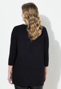 Schwarzes Oberteil mit Dreiviertelärmel, rundem Halsausschnitt und einem längeren Saum hinten. Glatte Textur und minimalistisches Design, keine sichtbaren Muster.