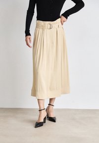 Jupe midi plissée beige avec une large ceinture dotée d'une boucle circulaire, assortie à des escarpins noirs à bout pointu et à brides de cheville.