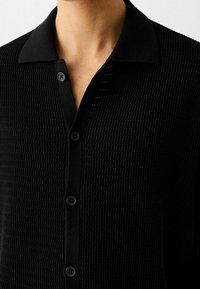Zwart, gestructureerd brei shirt met een kraag, zes knopen aan de voorkant en een patroon van verhoogde vierkanten.