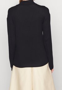 Maglietta nera a maniche lunghe con colletto alto. Realizzata in un tessuto liscio, presenta un design aderente e un orlo curvo. Lo sfondo è neutro.