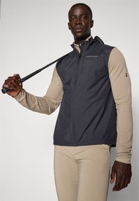 Ermeløs mørkegrå vest med lett design, glidelås og glatt tekstur, båret over en langermet beige topp. Golfklubb i hånden.