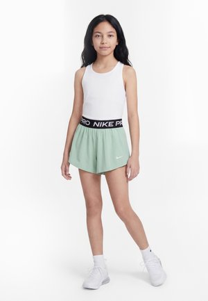Young girl standing, wearing white tank top, mint green Nike shorts with black waistband, white socks, and white sneakers on white background.