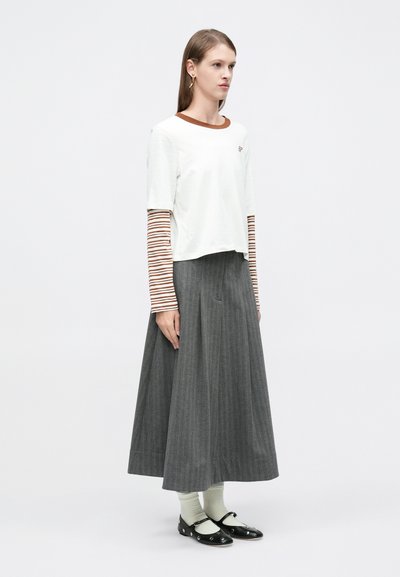 Λευκό κοντομάνικο τοπ με καφέ γιακά πάνω από μακριά ριγέ μανίκια, γκρι πλισέ μακρύ φούστα και μαύρα παπούτσια με διακοσμητικά στοιχεία.