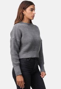 Grå kortad tröja med ett texturerat stickat mönster, ribbad nederkant och långa ärmar. I kombination med mörka jeans, som visar en enkel design.