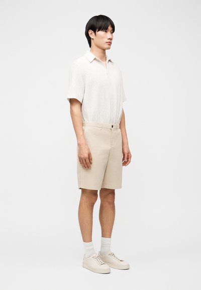 Νεαρός άνδρας που στέκεται, φοράει άσπρο κοντομάνικο πουκάμισο, μπεζ σορτς, λευκές κάλτσες και ανοιχτόχρωμα αθλητικά παπούτσια μπροστά σε λευκό φόντο.