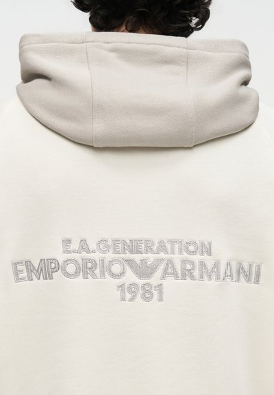 Άτομο με σκούρα, σγουρά μαλλιά που φοράει μπεζ φούτερ με κουκούλα με κεντημένο στο πίσω μέρος το κείμενο "E.A. Generation Emporio Armani 1981".