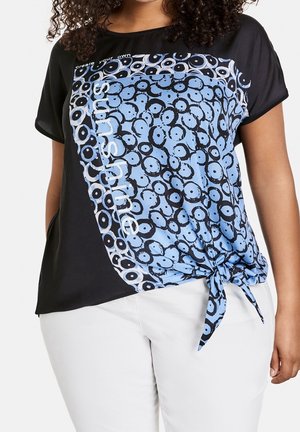 Camiseta negra de manga corta con un patrón circular azul y negro y texto "sunshine". Lado izquierdo anudado a la cintura. Textura suave.