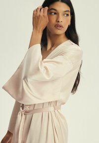 Jasnoróżowa satynowa sukienka z głębokim dekoltem w kształcie V, szerokimi rękawami i wiązaniem w talii. Gładka tekstura z pliskami na spódnicy.